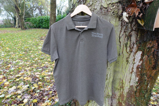 Green polo shirt in size Large hanging outdoors, showing the hand-stamped ‘The Earth Teaches’ print on the left chest
