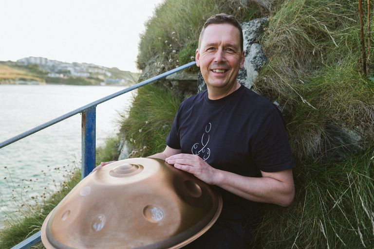 Man holding a handpan by a lakeside with greenery in the background