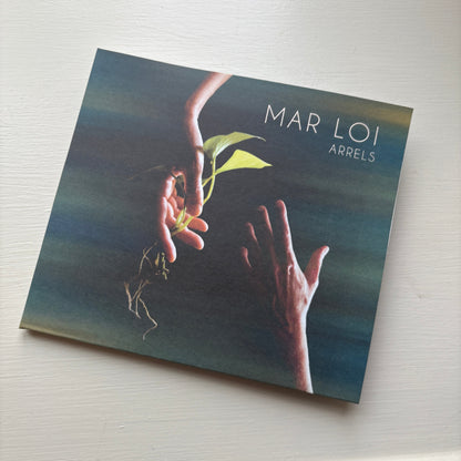 Mar Loi “Arrels” CD cover resting on a white surface, showing two hands reaching toward a small plant.